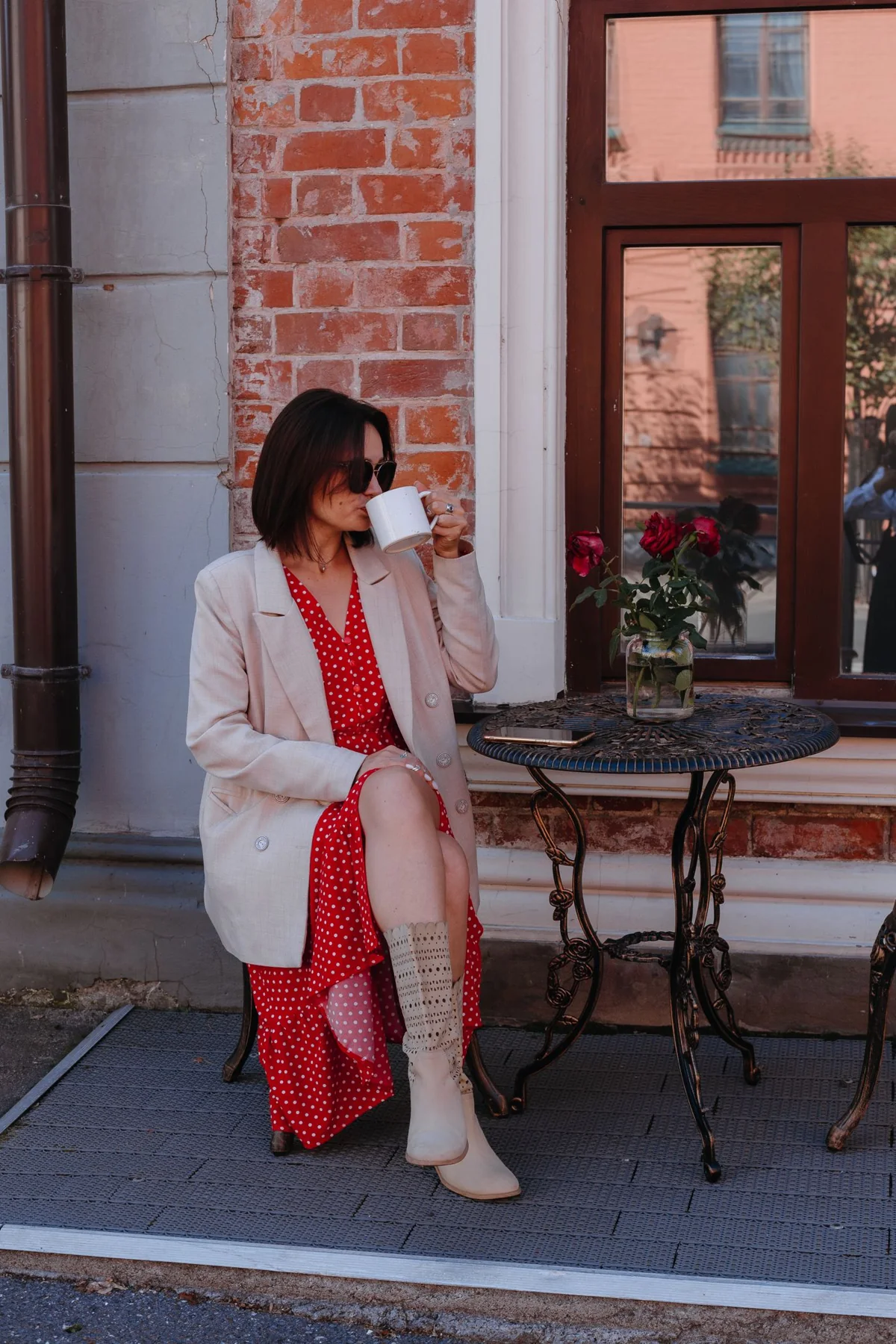 A woman in a light suit drinking from a cup at an outdoor cafe table.