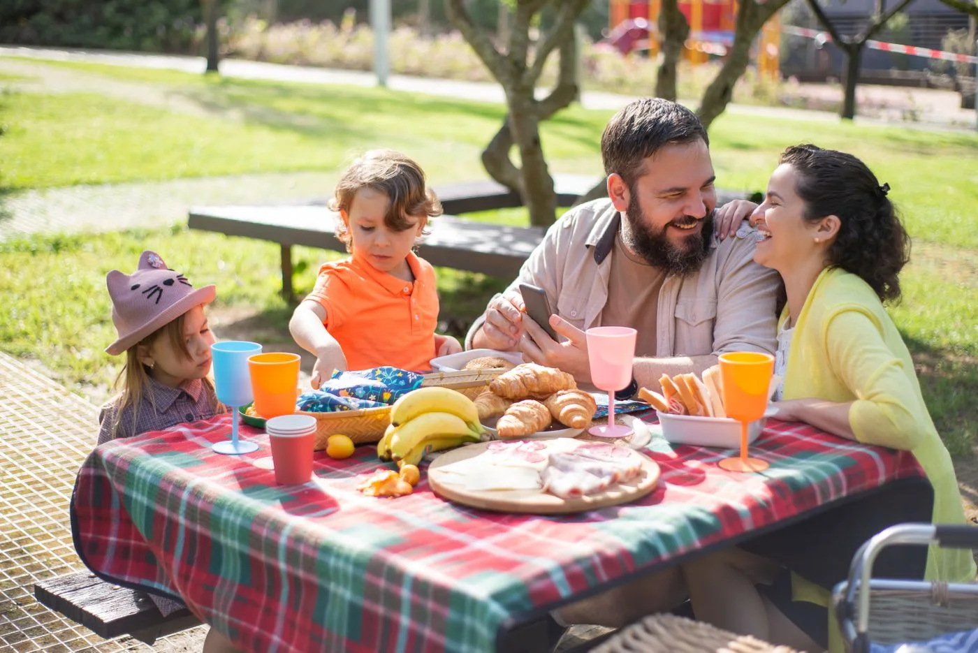 Adults and children sharing food around a checked outdoor picnic table.