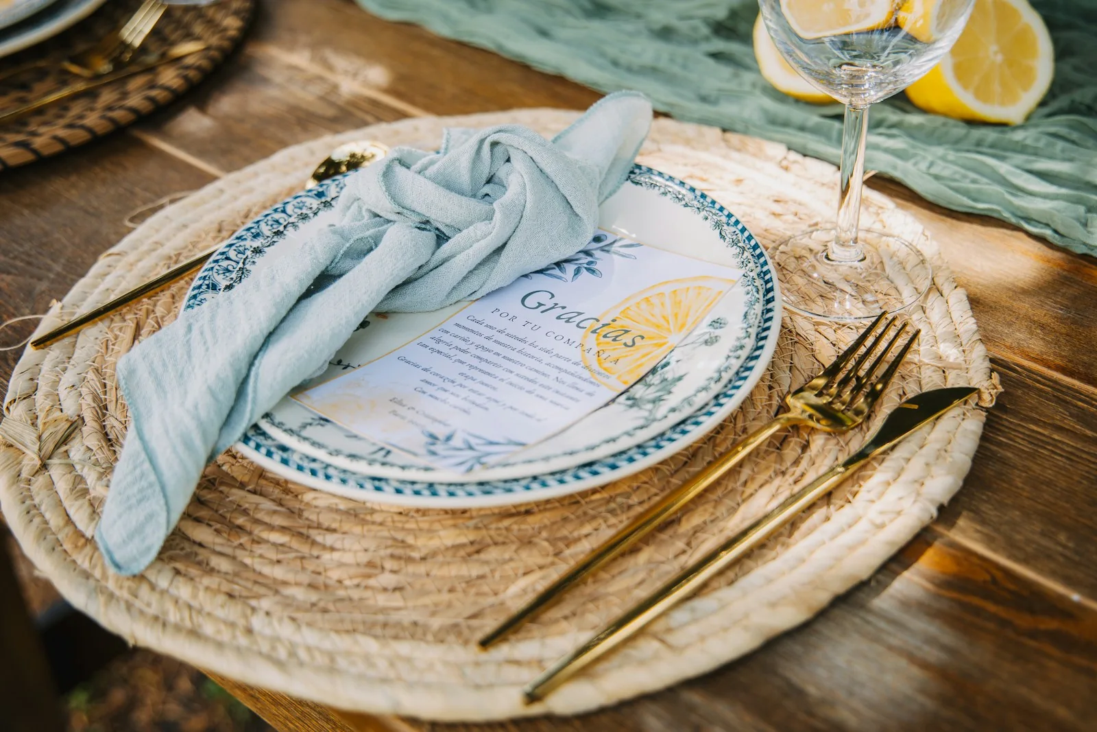 Patterned dinner plate with a pale cloth napkin, gold cutlery and a stemmed glass on a woven placemat