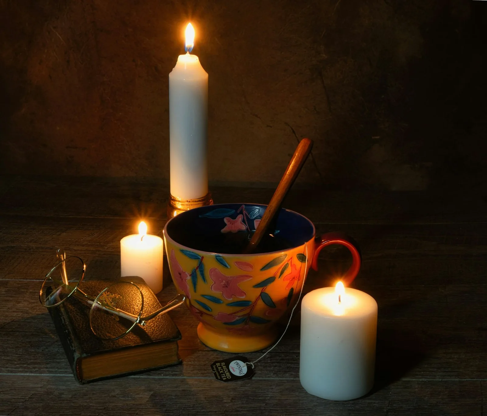 A colourful teacup beside lit candles, a small book and reading glasses.