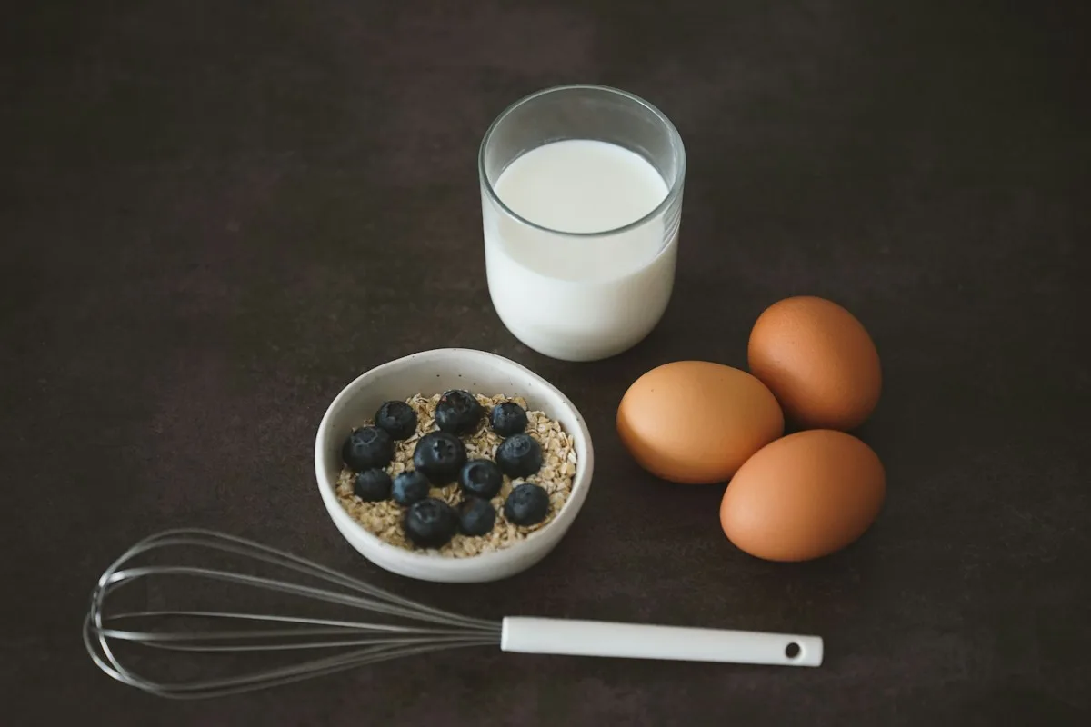 A bowl of blueberries beside eggs, a glass of milk and a whisk on a dark surface