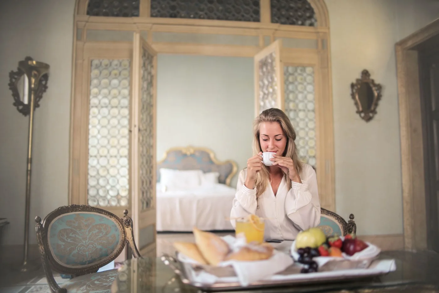 A woman drinking a hot beverage beside a breakfast tray in a hotel room.