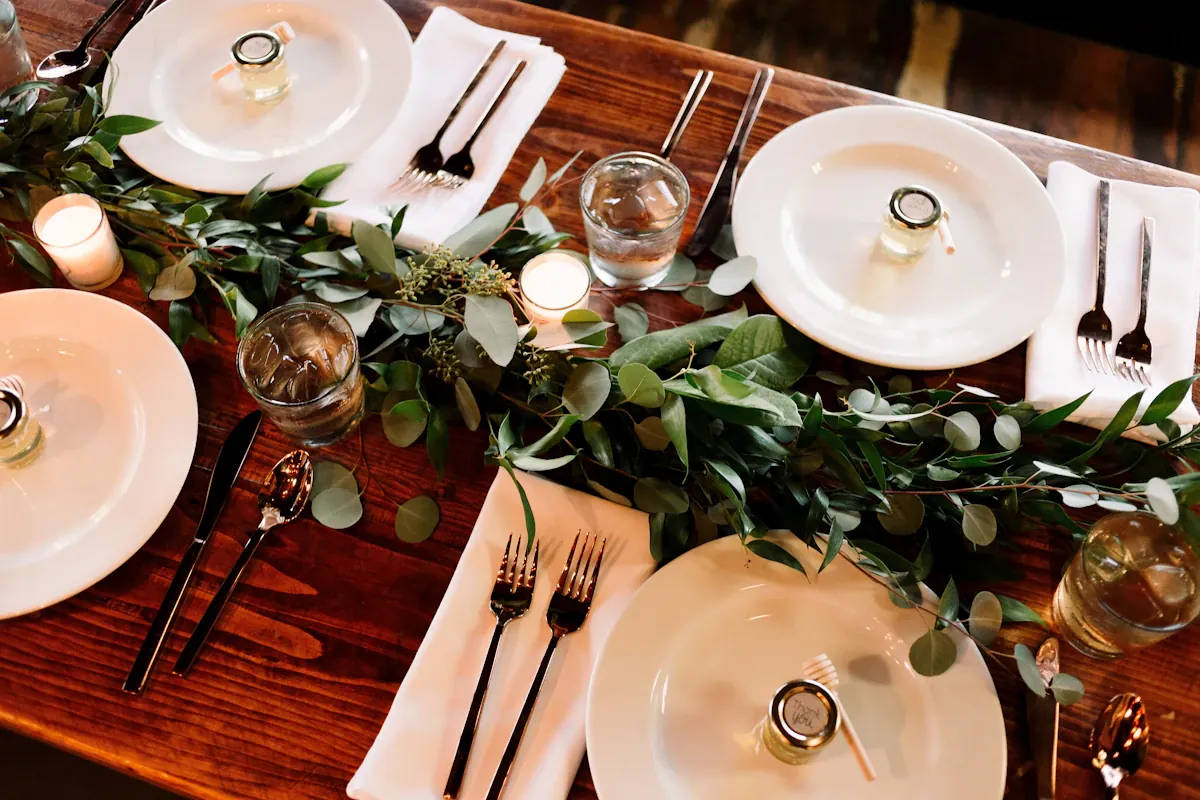 A dining table set with mixed plates, utensils and greenery